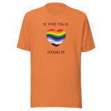 The image shows a unisex crew neck burnt orange t- shirt with a minimalist yet vibrant design. Centered on the front is a heart-shaped graphic filled with horizontal stripes in the colors of the progressive Pride flag, symbolizing inclusivity and diversity. Above the heart, the text reads "BE WHO YOU BE" and below it, "AROUND ME" both in sleek, black font. The design conveys an authenticity, making this t-shirt a wearable statement for equality and individuality.