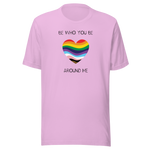 The image shows a unisex crew neck lilac t- shirt with a minimalist yet vibrant design. Centered on the front is a heart-shaped graphic filled with horizontal stripes in the colors of the progressive Pride flag, symbolizing inclusivity and diversity. Above the heart, the text reads "BE WHO YOU BE" and below it, "AROUND ME" both in sleek, black font. The design conveys an authenticity, making this t-shirt a wearable statement for equality and individuality.