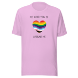 The image shows a unisex crew neck lilac t- shirt with a minimalist yet vibrant design. Centered on the front is a heart-shaped graphic filled with horizontal stripes in the colors of the progressive Pride flag, symbolizing inclusivity and diversity. Above the heart, the text reads "BE WHO YOU BE" and below it, "AROUND ME" both in sleek, black font. The design conveys an authenticity, making this t-shirt a wearable statement for equality and individuality.