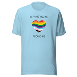 The image shows a unisex crew neck ocean blue t- shirt with a minimalist yet vibrant design. Centered on the front is a heart-shaped graphic filled with horizontal stripes in the colors of the progressive Pride flag, symbolizing inclusivity and diversity. Above the heart, the text reads "BE WHO YOU BE" and below it, "AROUND ME" both in sleek, black font. The design conveys an authenticity, making this t-shirt a wearable statement for equality and individuality.