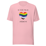 The image shows a unisex crew neck pink t- shirt with a minimalist yet vibrant design. Centered on the front is a heart-shaped graphic filled with horizontal stripes in the colors of the progressive Pride flag, symbolizing inclusivity and diversity. Above the heart, the text reads "BE WHO YOU BE" and below it, "AROUND ME" both in sleek, black font. The design conveys an authenticity, making this t-shirt a wearable statement for equality and individuality.