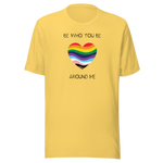 The image shows a unisex crew neck yellow t- shirt with a minimalist yet vibrant design. Centered on the front is a heart-shaped graphic filled with horizontal stripes in the colors of the progressive Pride flag, symbolizing inclusivity and diversity. Above the heart, the text reads "BE WHO YOU BE" and below it, "AROUND ME" both in sleek, black font. The design conveys an authenticity, making this t-shirt a wearable statement for equality and individuality.